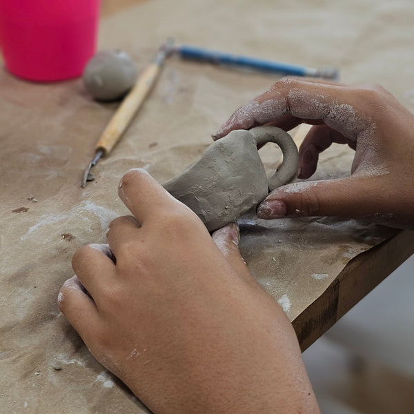 Hands working with clay on a pottery wheel, with a pink cup in the foreground.