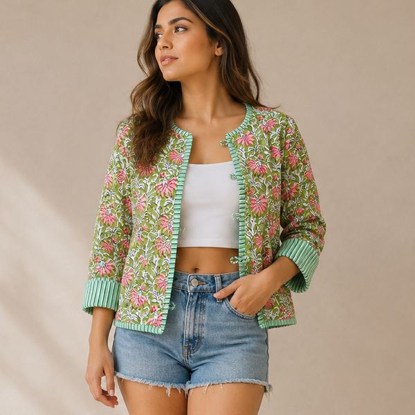 A woman models a green floral patterned knit cardigan with green ribbed cuffs and collar, styled with denim shorts, a white crop top, and white sneakers against a beige studio backdrop with window shadow patterns.
