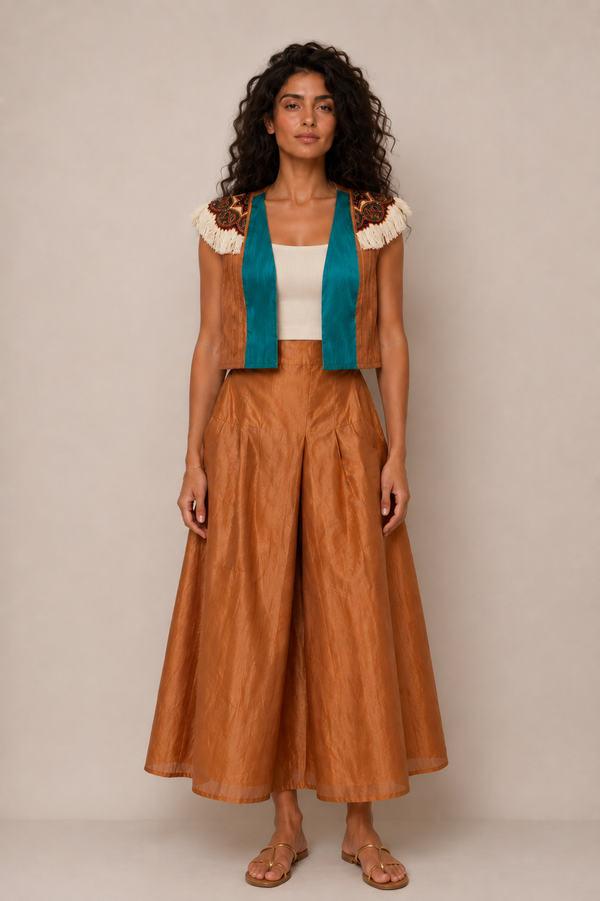 A woman stands against a plain beige studio background wearing a coordinated outfit: a rust-brown sleeveless vest with cream and brown embroidered shoulders, a teal inner layer, a white top, matching rust-brown wide-leg flared pants, and tan sandals.