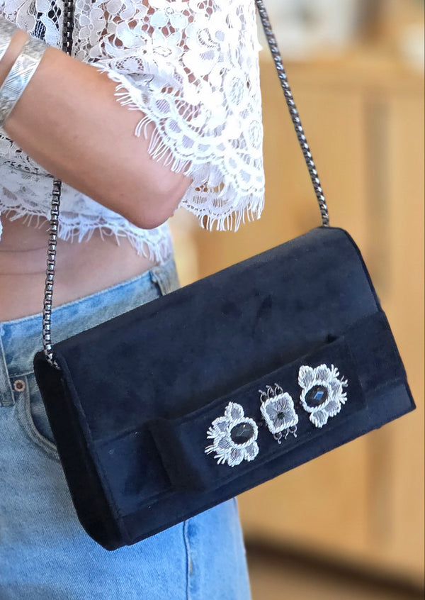 Person holding a black handbag with floral embroidery, wearing a white lace dress and silver bracelet.