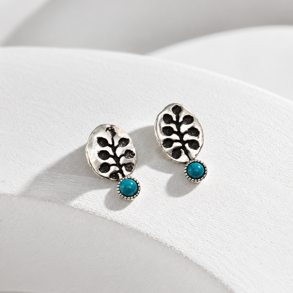 A pair of silver-toned drop earrings with an openwork leaf design and a small round turquoise blue stone at the bottom, displayed on a white curved surface.
