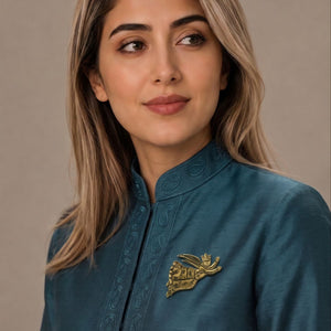 A woman with blonde-highlighted hair wearing a teal blue embroidered kurta with a gold metallic chest pin, photographed against a neutral beige background.