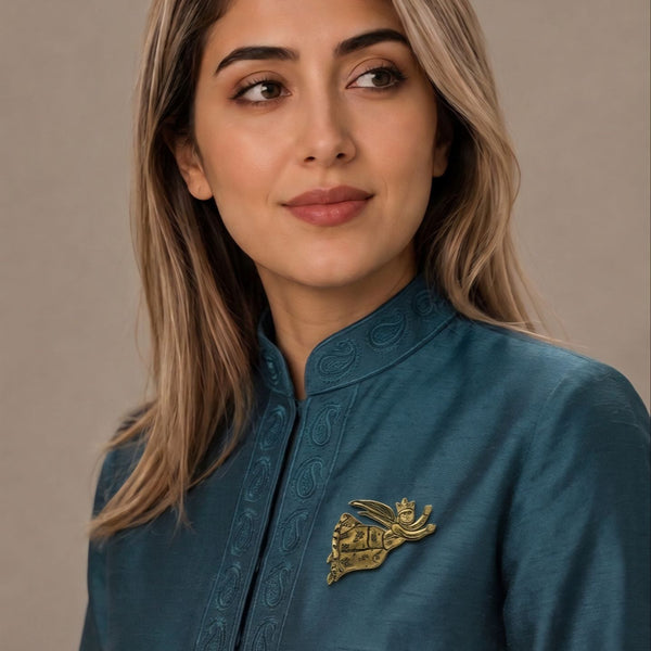 A woman with blonde-highlighted hair wearing a teal blue embroidered kurta with a gold metallic chest pin, photographed against a neutral beige background.