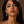 A close-up portrait of a woman with dark hair wearing silver teardrop dangle earrings with clear stones, paired with a burgundy satin spaghetti strap top, set against a solid brown background.