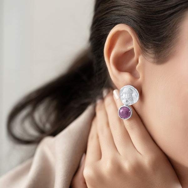 A close-up lifestyle shot of a model wearing a two-part earring. The earring features a textured silver disc on top with a small, round purple gemstone dangling below.