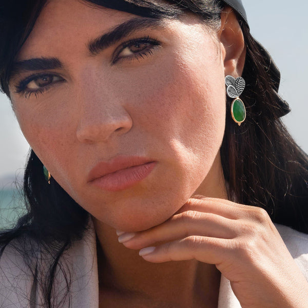 A close-up portrait of a woman wearing a silver-toned textured drop earring with a green teardrop-shaped stone, set against a bright outdoor background.