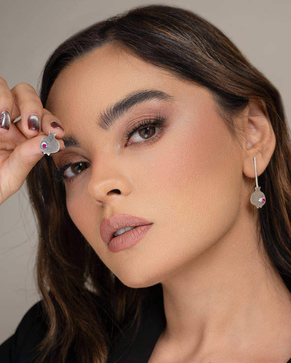 A close-up of a woman wearing a silver dangling earring with a small pink gemstone, holding the matching earring near her face with her hand.