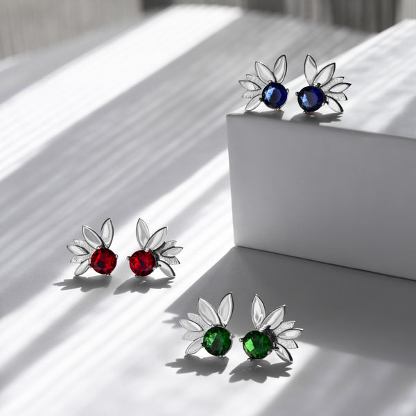 Three pairs of butterfly-shaped stud earrings displayed on a white geometric block against a sunlit white background. The top-right pair has blue oval stones, the bottom-right pair has green oval stones, and the left pair has red oval stones.