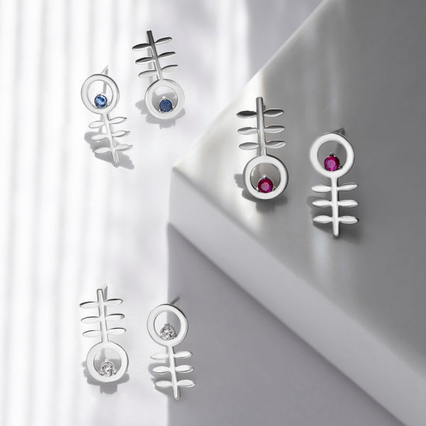 Five silver stud earrings with stylized fishhook or abstract geometric designs, some featuring small blue, pink, and clear gemstones, displayed on a white geometric surface with soft window shadows.