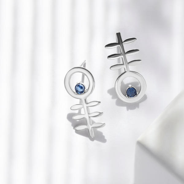 Five silver stud earrings with stylized fishhook or abstract geometric designs, some featuring small blue, pink, and clear gemstones, displayed on a white geometric surface with soft window shadows.