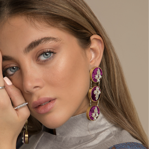 A close-up portrait of a woman wearing large oval drop earrings with purple and white stones in gold-toned settings, along with silver rings, against a warm beige background.