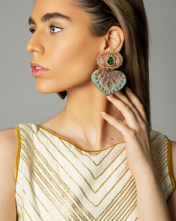 A close-up of a woman wearing a large, ornate double-drop statement earring. The earring features a gold-toned top disc with pink and green accents, and a large, textured heart-shaped drop in light blue and gold tones.