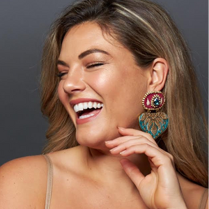 A woman with long brown hair is shown in a close-up portrait, smiling and wearing a large ornate statement earring with turquoise, red, and gold details against a dark gray background.