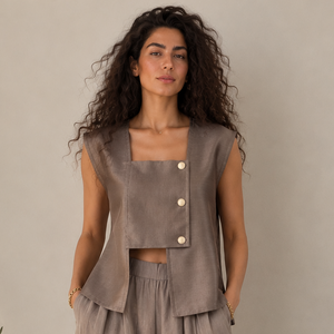 A woman modeling a taupe two-piece linen set consisting of a sleeveless cropped vest with buttons and matching wide-leg harem pants, standing against a neutral beige wall.