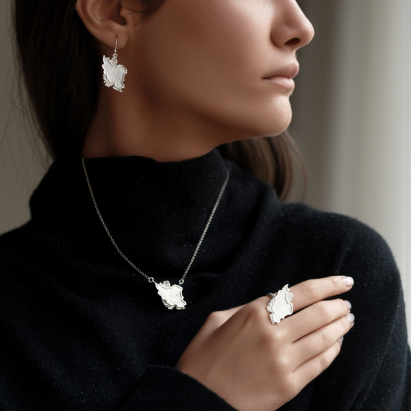A woman in a black turtleneck sweater models a matching jewelry set featuring a silver-toned necklace, ring, and earring, each with a textured, organic white leaf or flower-like pendant.
