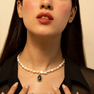A close-up portrait of a woman with dark hair wearing a pearl necklace with a green teardrop pendant, set against a plain beige background.