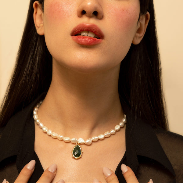 A close-up portrait of a woman with dark hair wearing a pearl necklace with a green teardrop pendant, set against a plain beige background.