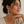 A woman wearing a statement dangle earring with a coral orange stone, silver-toned geometric detailing, and a white teardrop pearl, posed against a neutral beige background.