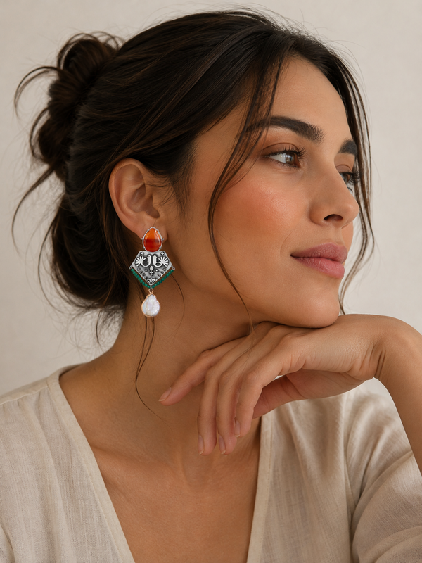 A woman wearing a statement dangle earring with a coral orange stone, silver-toned geometric detailing, and a white teardrop pearl, posed against a neutral beige background.