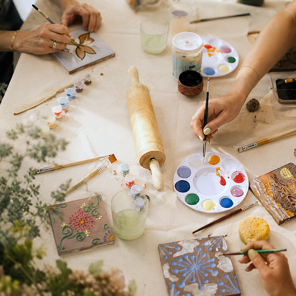 A group of people painting on small decorative tiles at a table covered with paint palettes, brushes, water, and floral decor.