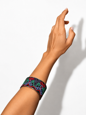 A close-up of a person's arm and hand wearing a wide black cuff bracelet with colorful embroidered swirl patterns in red, green, blue, and purple, set against a plain white background with a soft shadow.