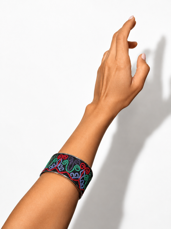 A close-up of a person's arm and hand wearing a wide black cuff bracelet with colorful embroidered swirl patterns in red, green, blue, and purple, set against a plain white background with a soft shadow.