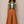 A woman stands against a plain beige studio background wearing a coordinated outfit: a rust-brown sleeveless vest with cream and brown embroidered shoulders, a teal inner layer, a white top, matching rust-brown wide-leg flared pants, and tan sandals.
