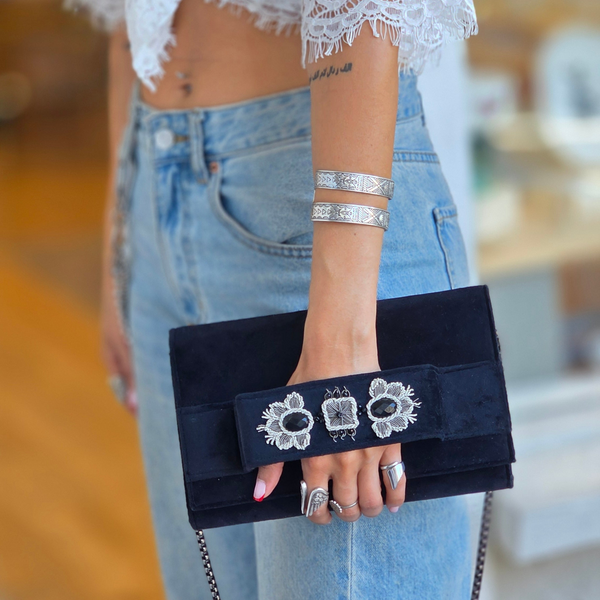 Person holding a black clutch with floral design, wearing lace sleeves and denim jeans.