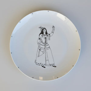 A white ceramic plate featuring a black line art illustration of a Qajar woman in traditional dress, placed on a textured gray concrete surface.