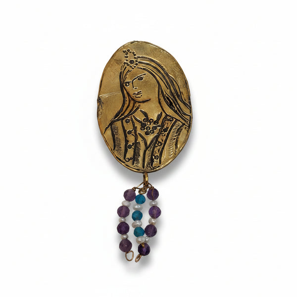 A vintage-style oval gold-toned pendant or brooch featuring an embossed portrait of a  Qajar woman, with dangling beaded strands in purple, blue, and white below, shown on a light textured background.