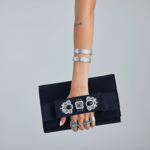 Person holding a black clutch with floral design, wearing lace sleeves and denim jeans.
