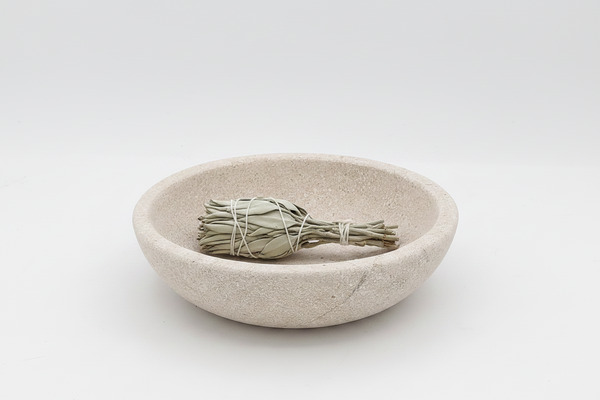 A round, shallow stone vessel sink or incense holder in a light beige speckled texture, featuring a small gold metal  holder in the center, set against a seamless light gray background.