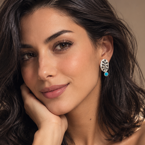 A woman with dark wavy hair poses in a close-up portrait, wearing a delicate silver bracelet with a round turquoise cabochon and a matching silver drop earring with a floral filigree design and a small turquoise accent.