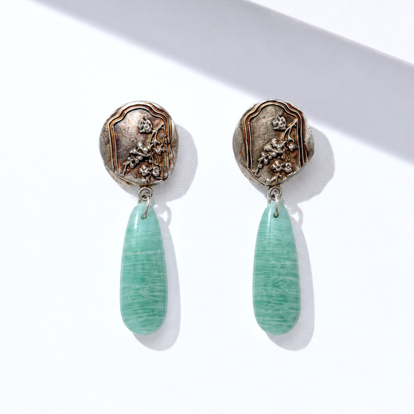 A pair of drop silver earrings with textured bronze-toned circular studs and dangling mint green teardrop-shaped beads, displayed on a clean white surface with a diagonal shadow.