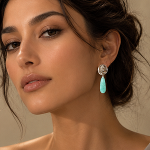 A close-up portrait of a woman wearing a dangling earring with a clear faceted top and a amazonite teardrop drop, against a warm beige background.