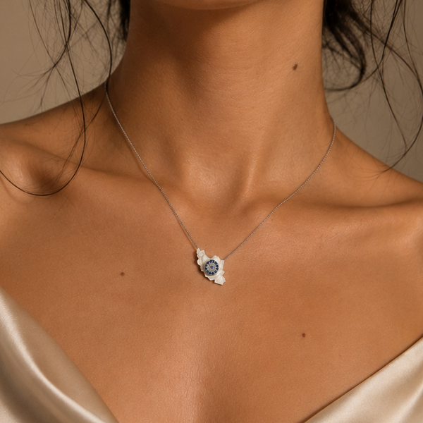 A close-up of a woman wearing a delicate silver Iran map necklace with a small evil eye pendant, styled with a cream satin camisole against a warm beige background.